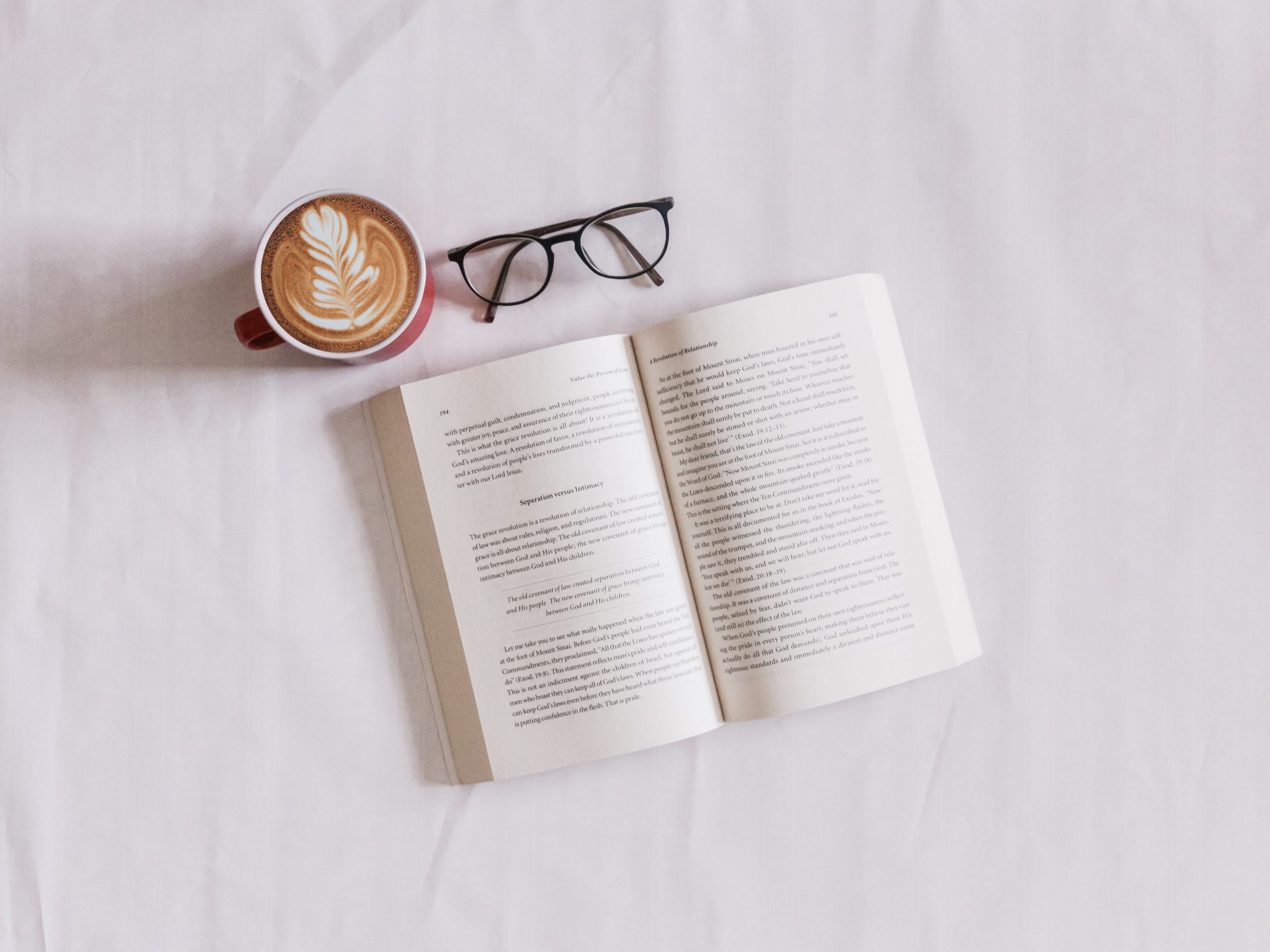 Book lying on a white sheet next to a pair of glasses and a cup of coffee