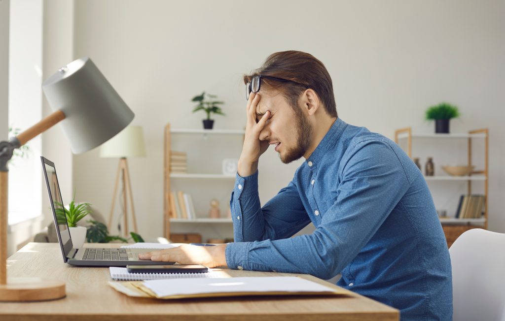 Tired stressed man sitting at desk with laptop and facepalming to a mistake he's made