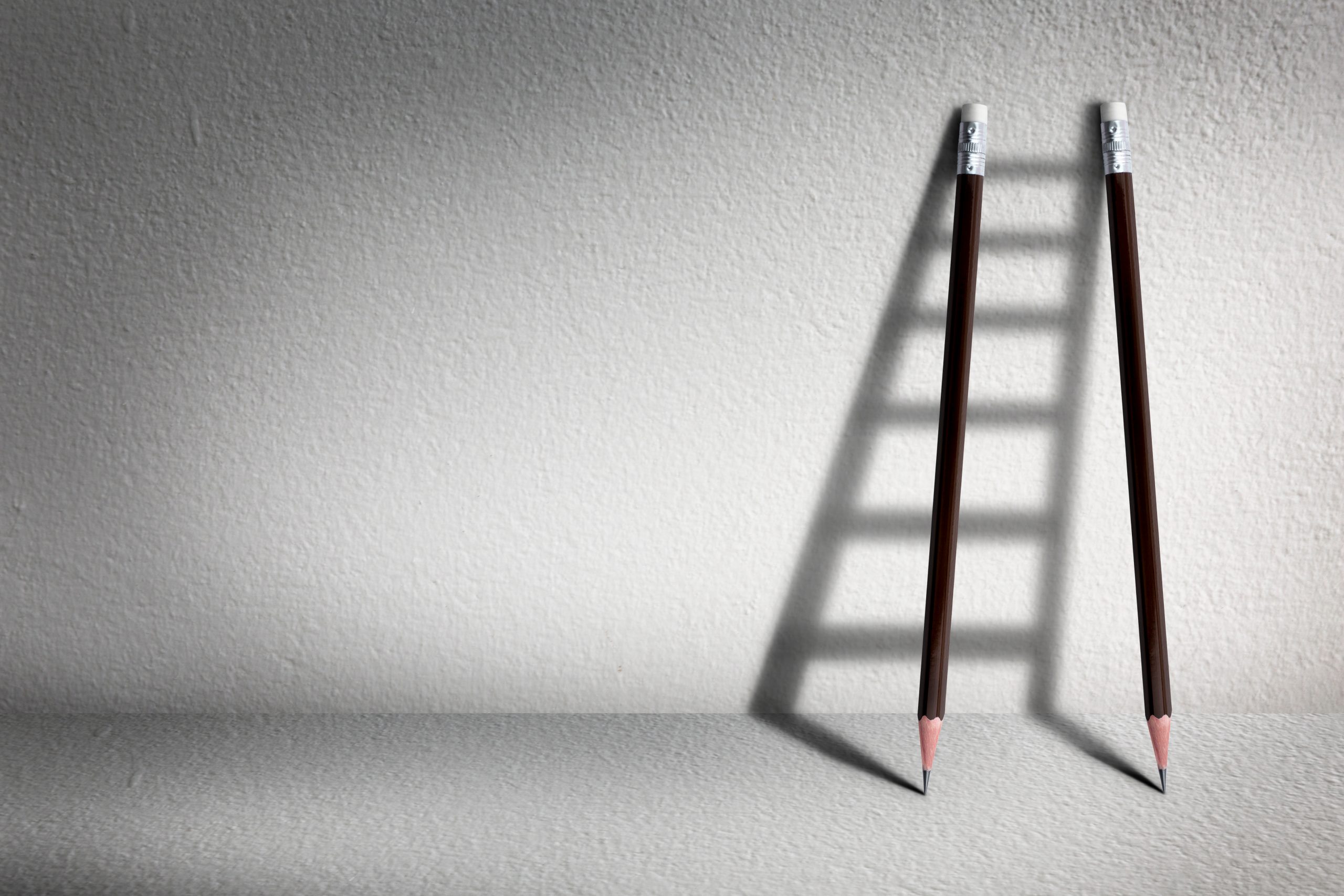 Two pencils lay next to a wall casting the shadow of a ladder