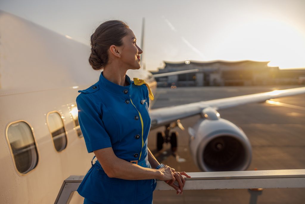 Thoughtful air stewardess in blue uniform looking away, standing outdoors at the sunset. Commercial airplane near terminal in an airport in the background
