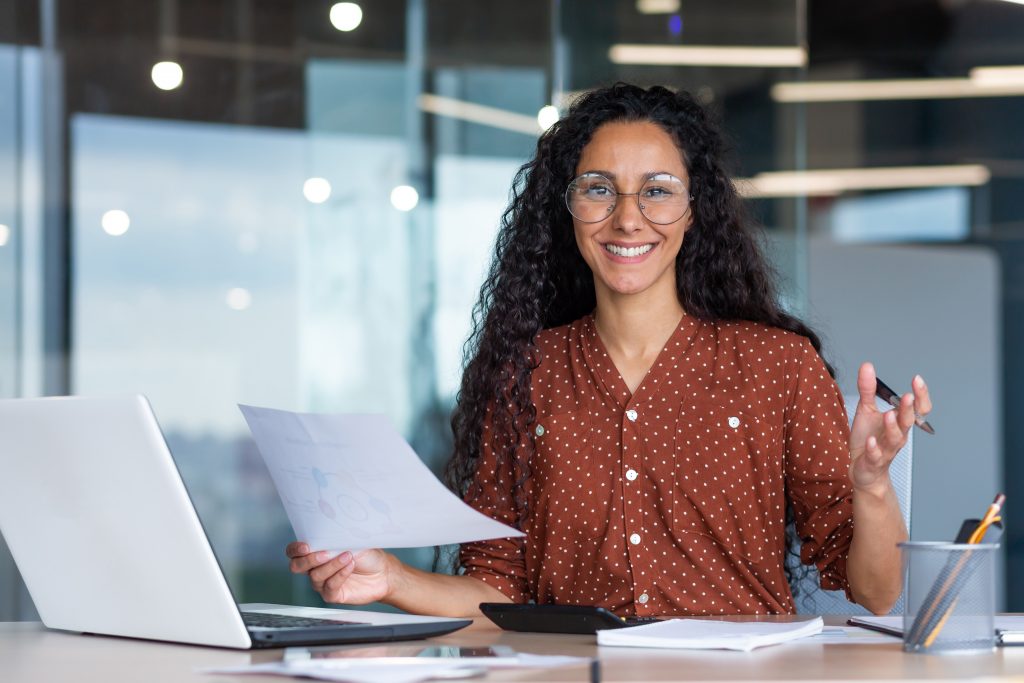 Portrait of happy and successful hispanic woman, businesswoman smiling and looking at camera holding contracts and invoices, working inside office with laptop on paper work