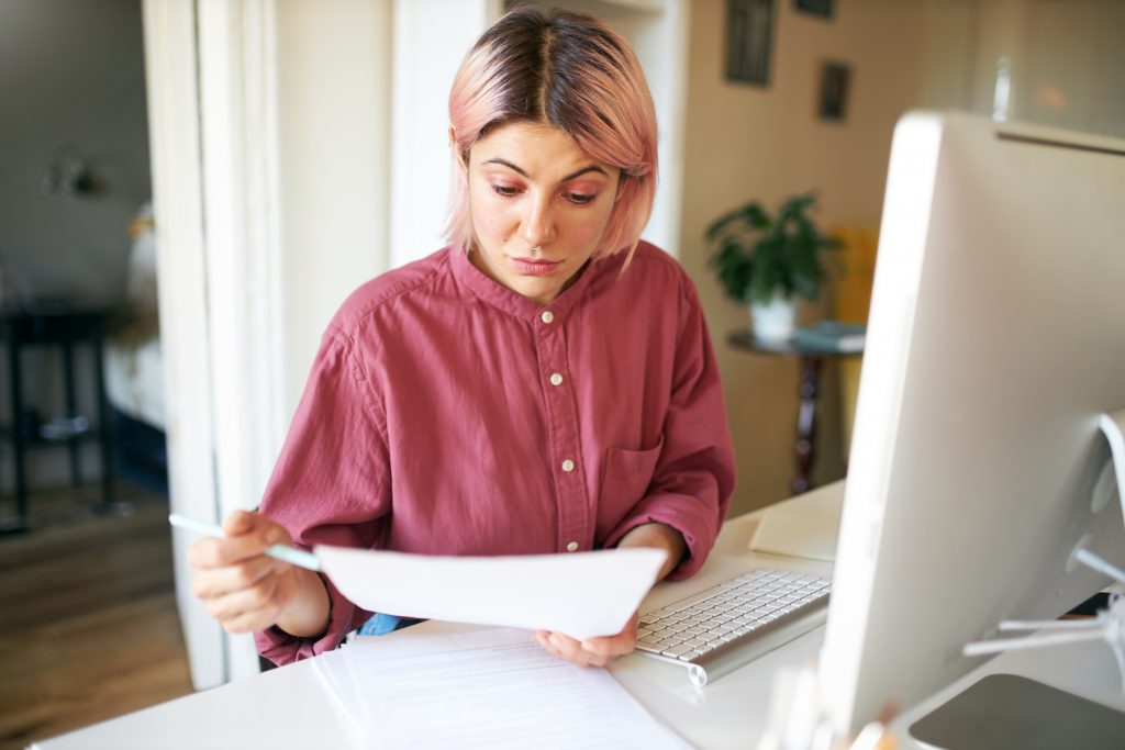 Portrait of concentrated young female manager working distantly from home sitting at desk with papers in her hands, studying agreement having serious facial expression, using desktop computer